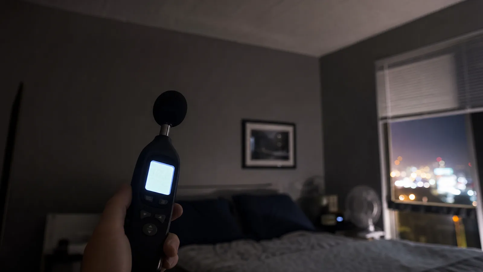 A handheld digital sound level meter glowing in a dark apartment bedroom