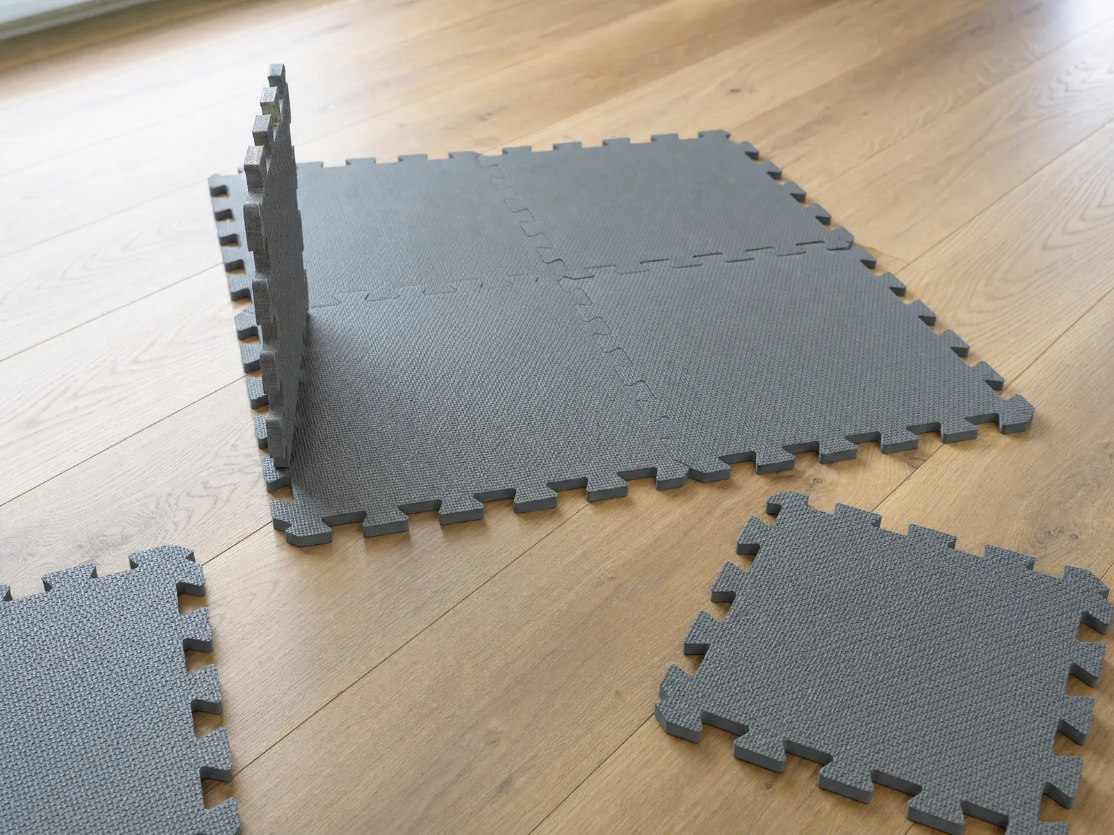 Grey interlocking foam puzzle tiles partly assembled on pale oak apartment flooring, one tile propped on its edge to show how thin it is